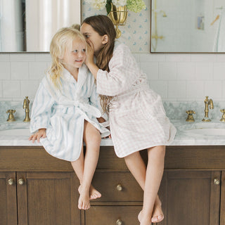 Two young girls sitting on a bathroom vanity, wearing light blue and pale pink checkered bathrobes. One girl is whispering to the other, who is smiling.