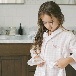 A young girl with long brown hair looks down while wearing a white and light pink gingham check pajama shirt with 'Mila' embroidered on the chest, standing in a bright bathroom.
