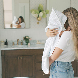 A woman in a white t-shirt and light blue jeans holds a baby wrapped in a white and light blue checkered blanket towel in a bright, modern bathroom.
