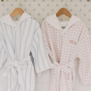 Two hooded bathrobes hanging on wooden hangers against patterned wallpaper. The left robe is white with light blue vertical stripes and a white hood.