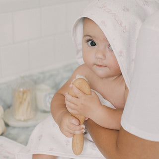 A baby wrapped in a white towel with a matching hood sits on an adult's lap, holding a wooden baby teething toy. The baby has a pink embroidered name on the hood, and the background shows a softly blurred bathroom setting.