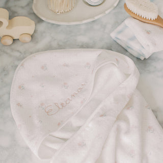 A white baby hooded towel with delicate pink floral embroidery and the name embroidered in pink script, styled on a marble surface with baby accessories.
