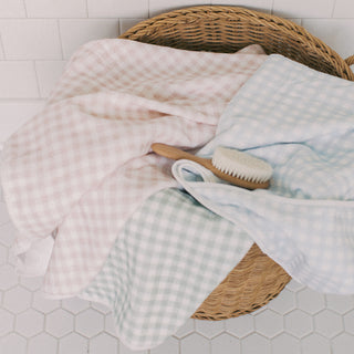 Three soft gingham towels in pale pink, light blue, and pale green are draped over a woven wicker basket, with a small wooden-handled brush resting on top, against a white tiled wall and floor.