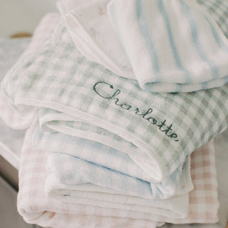 A stack of folded towels in soft pastel colors, including white, pale blue, and pale pink gingham and striped patterns. One cloth is embroidered with the name 'Charlotte' in dark green script.