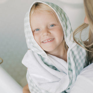 A smiling young child wrapped in a soft white towel with a pale green gingham hood, held by an adult in a bright, softly lit setting.