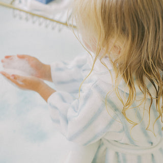 A child with wet blonde hair wearing a white and light blue striped bathrobe, seen from behind as they splash water in a bathtub.