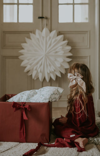 Child in a red dress with a large bow sitting next to a decorative box in a room with large windows.