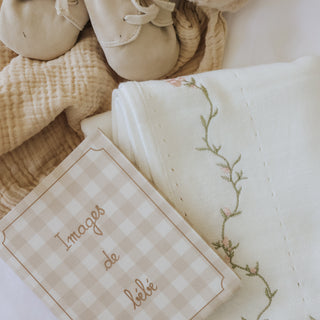 A styled flat-lay of baby textiles and accessories on a soft white background, including a pair of cream baby shoes, a plush teddy bear, a folded embroidered white knit blanket and a gingham photo album 'Images de bebe'.