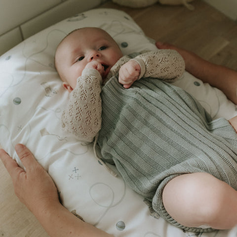 Baby in green outfit lying on a white cushion with hands gently holding the baby