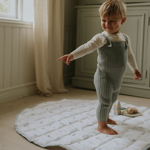 Child in a green striped outfit standing on a white mat in a room with light-colored walls and wooden floor.