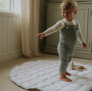 Child in a green striped outfit standing on a white mat in a room with light-colored walls and wooden floor.