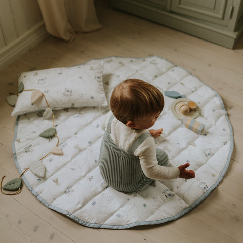 Child sitting on a round quilted mat with toys on a wooden floor.