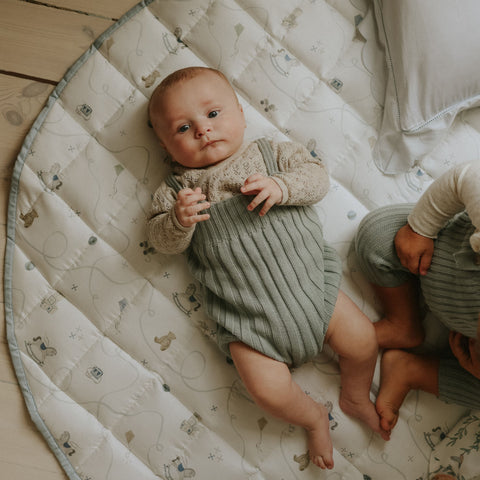 Baby lying on a patterned mat with another child partially visible
