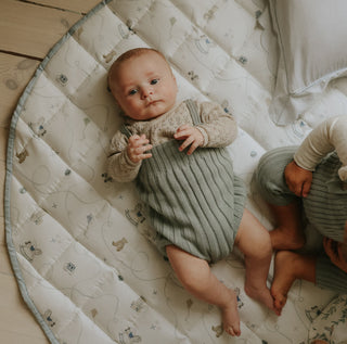 Baby lying on a patterned mat with another child partially visible