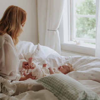 A woman in a white robe sits on a bed with white bedding, gently holding a baby dressed in a white outfit. A green-and-white checkered pillow is in the foreground, and a bright window with white curtains is visible in the background.