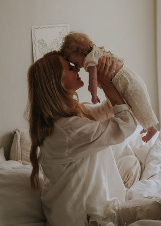A woman in a white robe holds a baby dressed in a matching white outfit while sitting on a bed in a softly lit bedroom.