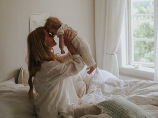 A woman in a white robe holds a baby dressed in a matching white outfit while sitting on a bed in a softly lit bedroom.