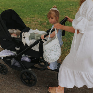 A woman in a white dress pushes a black stroller outdoors while a young child stands beside it holding a white green floral storage basket