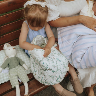 A young child sits on a wooden bench holding a quilted storage basket with green floral pattern. An adult woman in a white top and blue striped skirt sits beside them, and a plush bunny toy rests on a soft pillow to the left.