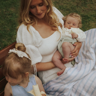 A woman in a white textured top holds a baby wrapped in a white and pale green blanket, with a young child in a light blue dress seated beside them on a bench outdoors.