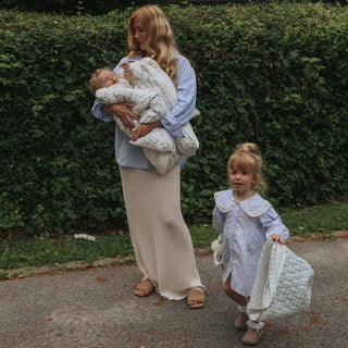 A woman outdoors carrying a baby wrapped in a light floral quilted blanket, with a young child beside her holding a matching light blue dress and quilted bag, set against a green hedge background.
