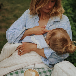 A woman in a light blue and white striped shirt holds a baby wrapped in a cream blanket outdoors. The woman is wearing a gold necklace and ring, and a green and white checkered blanket is visible underneath.