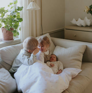 Three children on a couch in a cozy living room with a plant and lamp in the background.