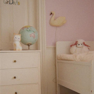 A soft pastel nursery scene with a white dresser, white single bed, small pink stool, globe, swan wall decor, and a plush toy on a pink wall.