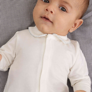 A close-up of a baby wearing a white long-sleeve sleep sack or romper with a Peter Pan collar, lying on a black and white gingham patterned blanket.