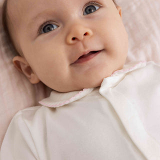A close-up of a smiling baby wearing a soft white  baby onesie with a pale pink ruffled collar, lying on a light pink blanket.