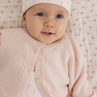 A close-up of a baby wearing a pale pink knitted cardigan and pink patterned hat, lying on a soft pink and white patterned blanket.