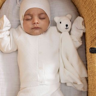 A white long-sleeve knit cardigan sweater with a button-up front on sleeping baby in bassinet.