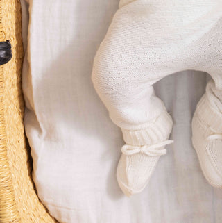 Close-up of a baby's legs wearing cream-colored knit booties with small bows, resting on a white muslin blanket.