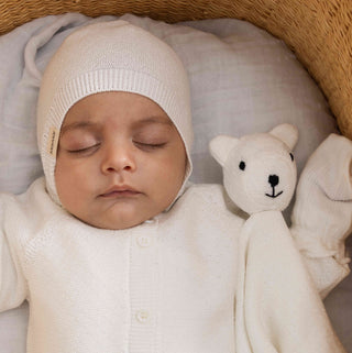 A sleeping newborn baby wearing a cream-colored knit bonnet and matching knit outfit, lying in a wicker bassinet next to a small plush teddy bear toy.