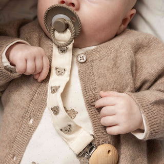 A close-up of a baby wearing a beige knit cardigan over a cream bodysuit, with a matching pacifier clip holding a pacifier. The pacifier clip strap is cream with small teddy bear prints, and the baby has a wooden button closure on the cardigan.