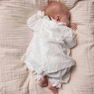 A baby sleeping on a textured beige muslin blanket, wearing a white long-sleeve blouse with ruffled details.