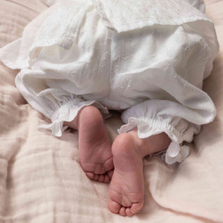 A close-up of a baby's bare feet peeking out from a white cotton pair of bloomer pants, resting on a light beige muslin fabric surface.