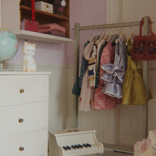 A styled children's room corner with a white dresser, open shelving, a clothing rack holding several dresses, a small toy piano, and a plush toy on a light floor against pale pink walls.