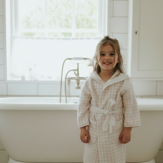 Child in a picnic gingham robe standing in a bathroom with a bathtub and window in the background.
