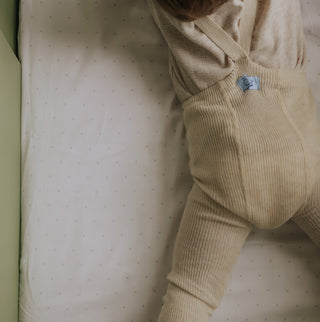 Beige overalls worn by a child on a white crib with subtle pattern