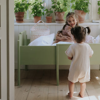 Two young children in a bedroom with a green bed and potted plants on a windowsill.
