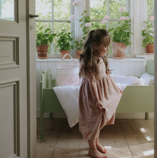 Young girl in a pink dress standing in a sunlit room with plants and a bed.