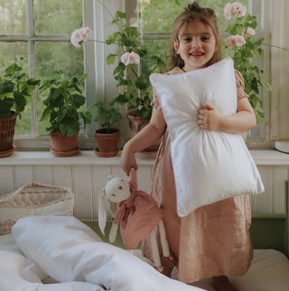 Child holding a pillow in a room with plants and a window.