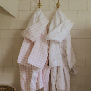 Two children's robes, one pink checkered and one white with floral patterns, hanging on a tiled wall.