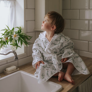 Child in a floral robe sitting on a bathroom counter by a sink.