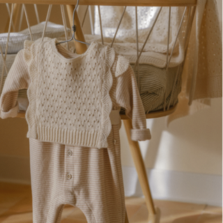 A baby knit two-piece outfit in warm cream and beige tones,  hanging on a wooden hanger in front of a wooden bassinet.