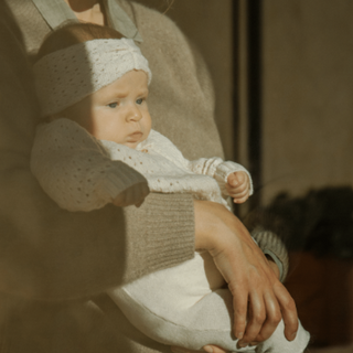 A baby wearing a white knit outfit and matching headband is held in the arms of an adult in warm, sunlit indoor lighting.