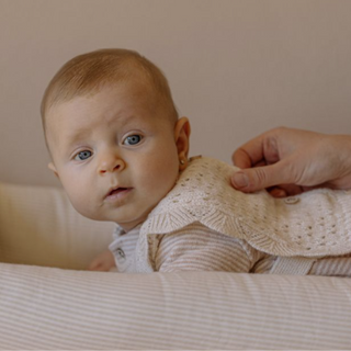 A close-up lifestyle shot of a baby wearing a cream-colored knit romper and matching knitted bib, with an adult hand gently holding the bib in place. The background is a soft, warm beige tone.