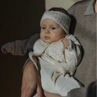 A baby wearing a cream-colored knit headband and matching knit outfit, held on a person's lap in a dimly lit indoor setting.