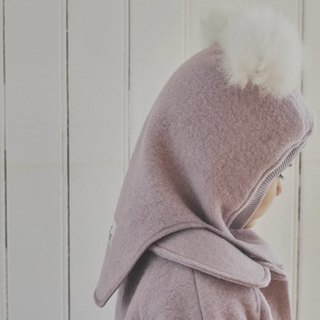 A child wearing a dusty pink fleece hooded coat with a fluffy white pom-pom on the hood, shown in profile against a white paneled wall.